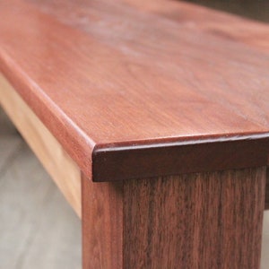 May include: Close-up of a wooden bench with a smooth, reddish-brown finish. The bench has a slightly rounded edge and a dark brown stain around the edge.