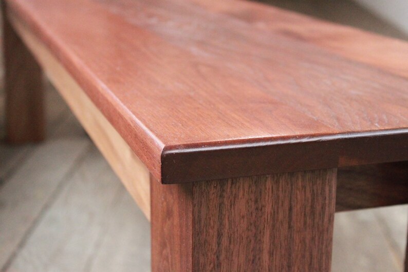 May include: Close-up of a wooden bench with a smooth, reddish-brown finish. The bench has a slightly rounded edge and a dark brown stain around the edge.