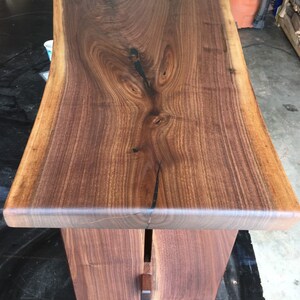 May include: A dark brown live edge walnut wood table with a natural finish. The table top is thick and has a unique grain pattern. The table legs are made of the same wood and are attached to the underside of the table top.