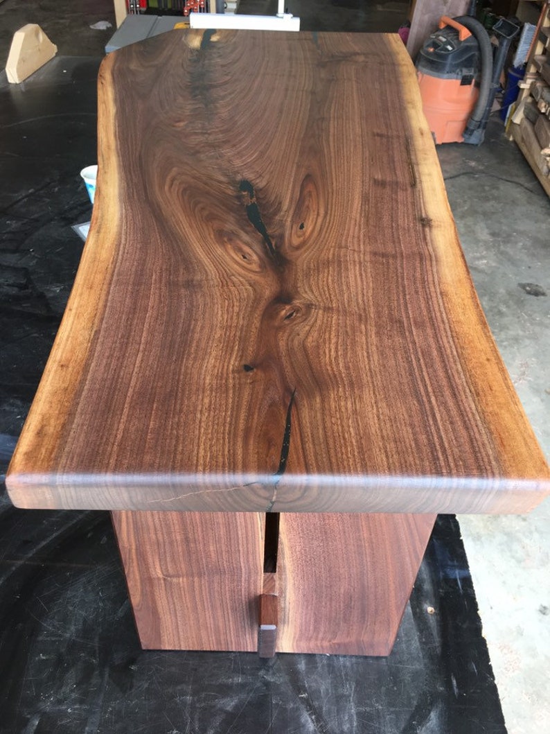 May include: A dark brown live edge walnut wood table with a natural finish. The table top is thick and has a unique grain pattern. The table legs are made of the same wood and are attached to the underside of the table top.