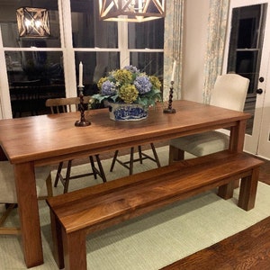 May include: A wooden dining table and bench set in a dining room. The table is made of dark wood and has a rectangular shape. The bench is also made of dark wood and has a rectangular shape. The table and bench are both in a room with a light green rug and hardwood floors. There is a floral arrangement in a blue and white vase on the table.