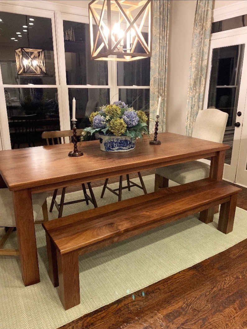 May include: A wooden dining table and bench set in a dining room. The table is made of dark wood and has a rectangular shape. The bench is also made of dark wood and has a rectangular shape. The table and bench are both in a room with a light green rug and hardwood floors. There is a floral arrangement in a blue and white vase on the table.