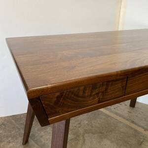 May include: A dark brown wooden desk featuring a smooth rectangular top with visible wood grain patterns and two integrated drawers with a seamless front design, supported by angled legs that extend outward from the main frame.