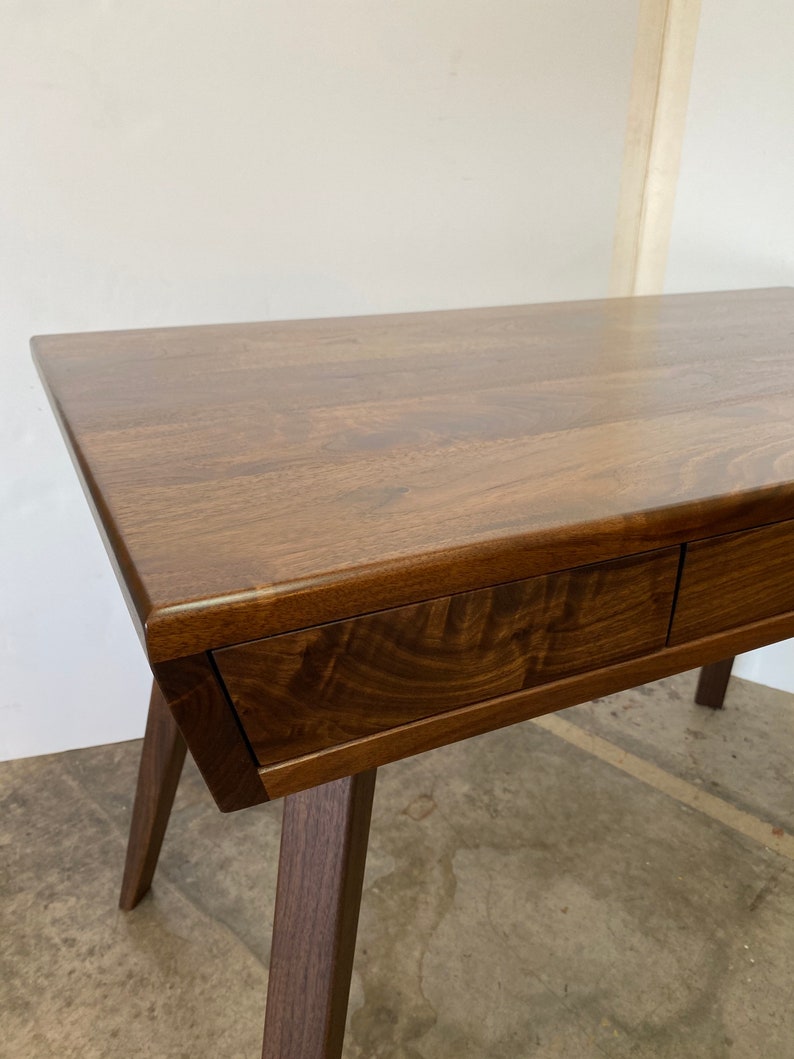 May include: A dark brown wooden desk featuring a smooth rectangular top with visible wood grain patterns and two integrated drawers with a seamless front design, supported by angled legs that extend outward from the main frame.