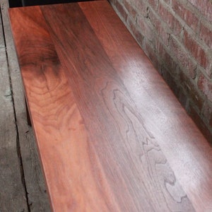May include: Close-up of a wooden bench top with a rich reddish-brown colour and a natural wood grain pattern. The wood is smooth and polished, with a slight sheen.