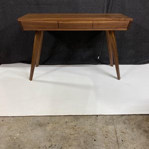 May include: A rectangular wooden desk featuring a rich brown grain pattern, three horizontal drawers aligned across the front, and four tapered legs that angle outward from the center to provide a stable base for this functional furniture piece.