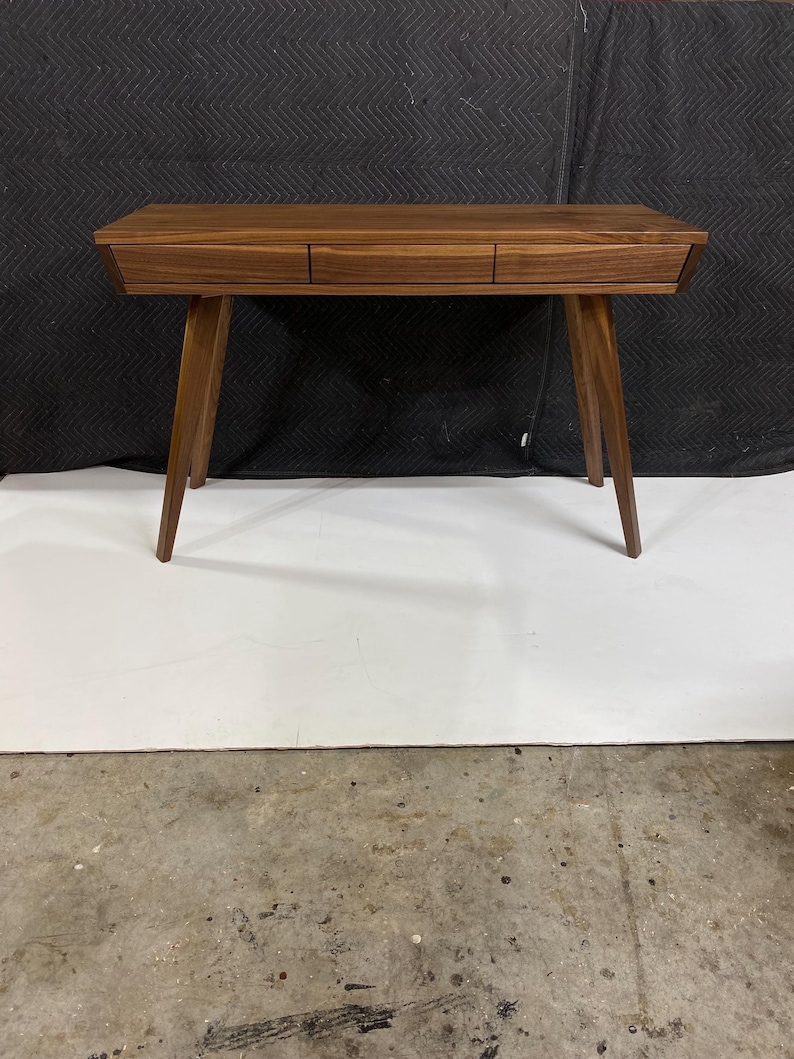 May include: A rectangular wooden desk featuring a rich brown grain pattern, three horizontal drawers aligned across the front, and four tapered legs that angle outward from the center to provide a stable base for this functional furniture piece.