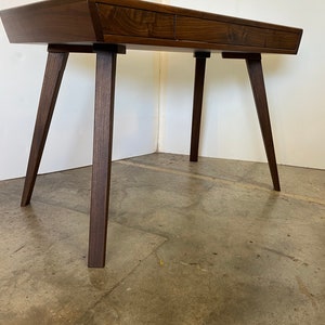 May include: A dark brown wooden desk featuring a smooth, rectangular top with three integrated drawers across the front. The desk is supported by four angled, tapered legs that extend outward from the corners, providing a stable base.