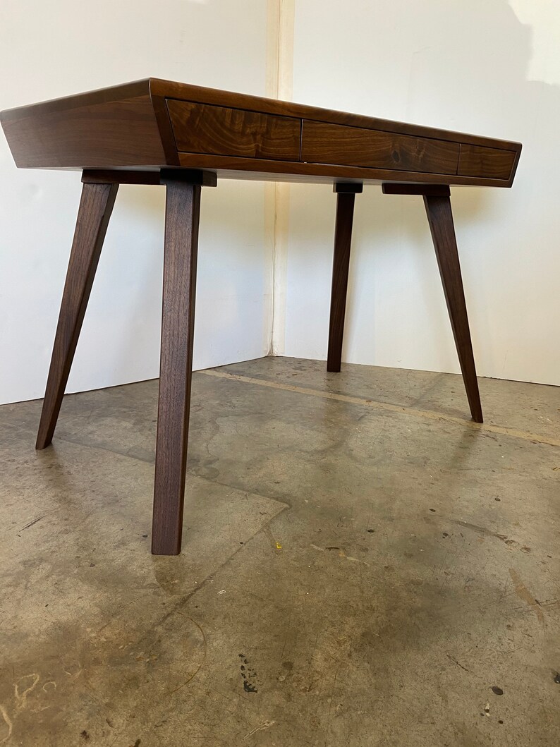 May include: A dark brown wooden desk featuring a smooth, rectangular top with three integrated drawers across the front. The desk is supported by four angled, tapered legs that extend outward from the corners, providing a stable base.