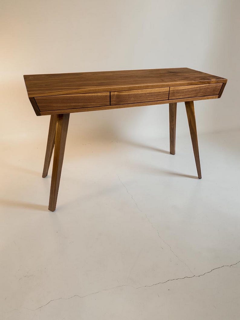 May include: A rectangular wooden desk featuring a rich brown grain pattern, three horizontal drawers aligned across the front, and four tapered legs that angle outward from the base to provide structural support for the flat tabletop surface.