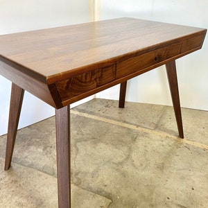 May include: A dark brown wooden desk featuring a rectangular top with a smooth grain finish. The desk includes two integrated drawers positioned horizontally along the front edge and is supported by four tapered, angled legs made of wood.