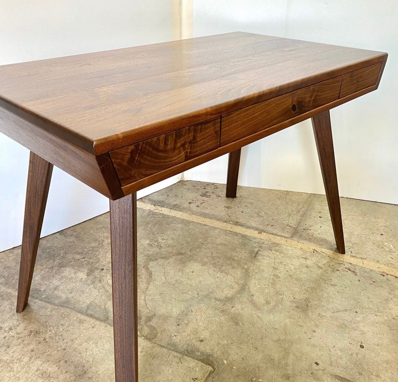 May include: A dark brown wooden desk featuring a rectangular top with a smooth grain finish. The desk includes two integrated drawers positioned horizontally along the front edge and is supported by four tapered, angled legs made of wood.