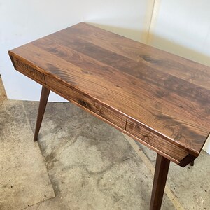 May include: A rectangular wooden desk featuring a dark brown grain pattern, a smooth flat top surface, and two integrated drawers along the front edge. The desk is supported by four tapered wooden legs angled outward from the base of the frame.