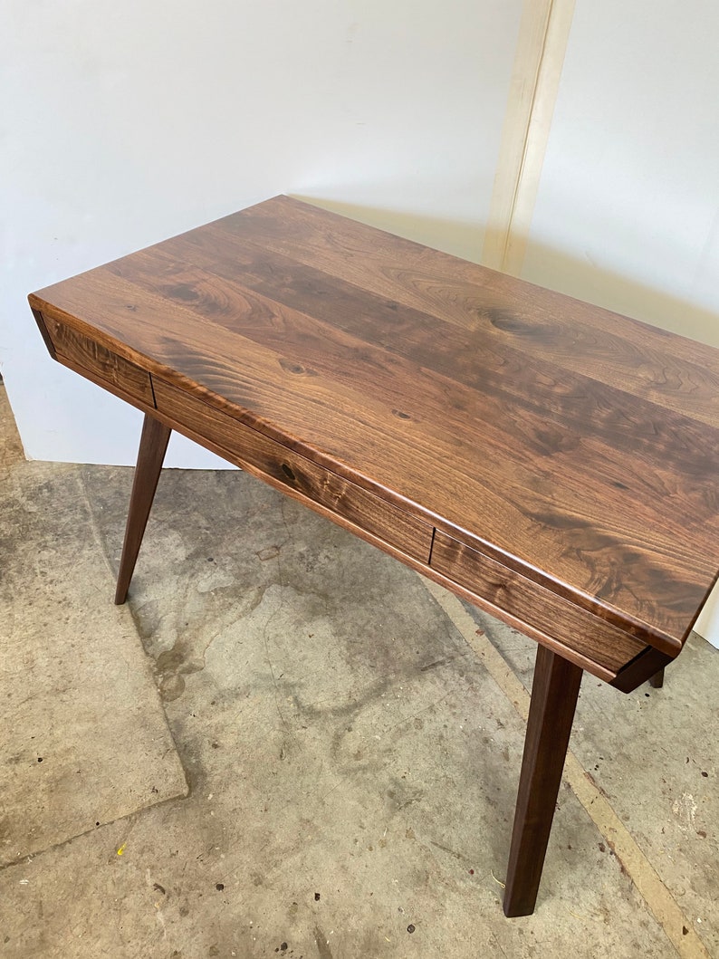 May include: A rectangular wooden desk featuring a dark brown grain pattern, a smooth flat top surface, and two integrated drawers along the front edge. The desk is supported by four tapered wooden legs angled outward from the base of the frame.