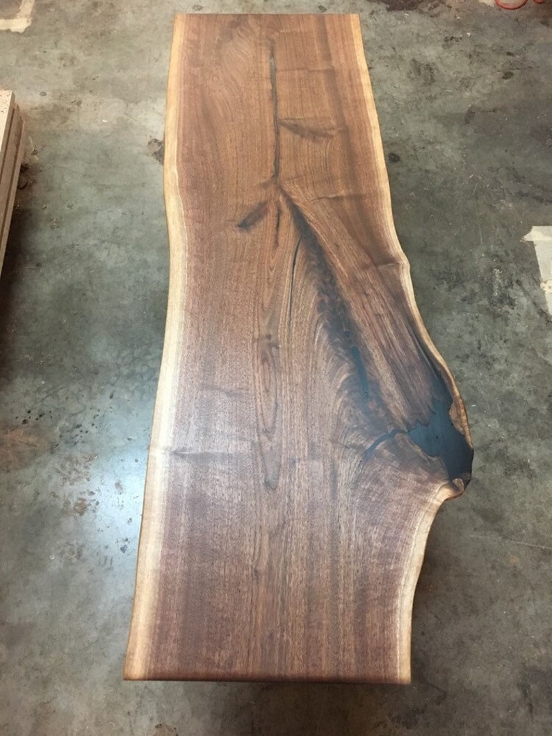 May include: A large, live edge slab of walnut wood with a natural, dark brown finish. The wood has a unique grain pattern and a black epoxy fill in a crack.
