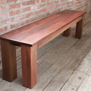 May include: A solid wood bench with a natural wood finish. The bench has a rectangular top and four legs. The bench is made of dark wood and has a smooth surface.