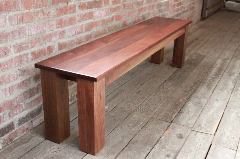 May include: A solid wood bench with a natural wood finish. The bench has a rectangular top and four legs. The bench is made of dark wood and has a smooth surface.