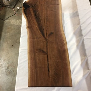 May include: A large, live edge slab of walnut wood with a natural, dark brown finish. The wood has a unique grain pattern and a live edge on both sides.