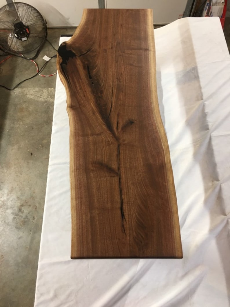 May include: A large, live edge slab of walnut wood with a natural, dark brown finish. The wood has a unique grain pattern and a live edge on both sides.