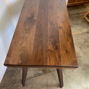 May include: A rectangular table crafted from dark brown wood with a visible natural grain pattern and knots. The table features a smooth, flat top surface and four sturdy, angled legs extending downward from the corners of the rectangular frame.