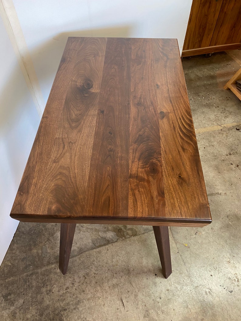 May include: A rectangular table crafted from dark brown wood with a visible natural grain pattern and knots. The table features a smooth, flat top surface and four sturdy, angled legs extending downward from the corners of the rectangular frame.