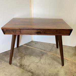May include: A dark brown wooden desk featuring a rectangular top with a visible wood grain pattern. The desk includes a single long central drawer flanked by two smaller side compartments, supported by four tapered legs angled outward.