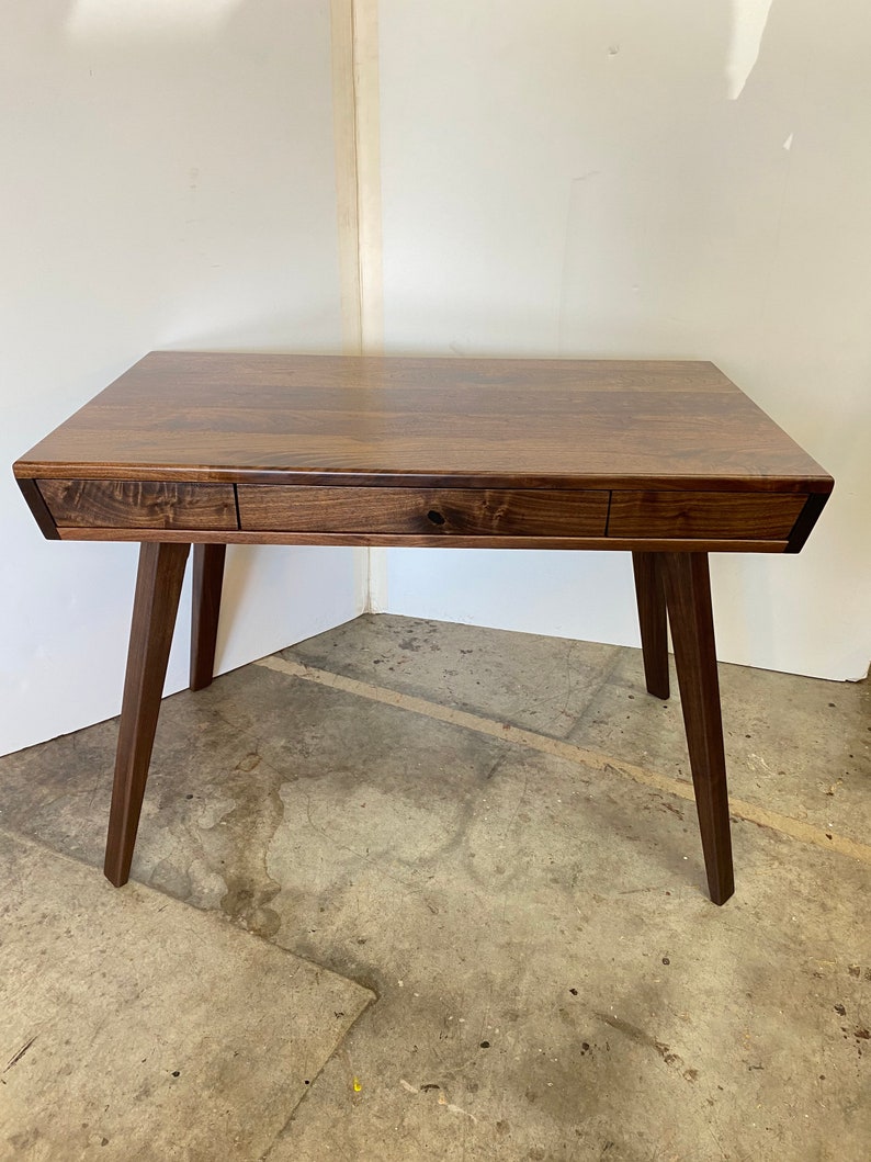 May include: A dark brown wooden desk featuring a rectangular top with a visible wood grain pattern. The desk includes a single long central drawer flanked by two smaller side compartments, supported by four tapered legs angled outward.