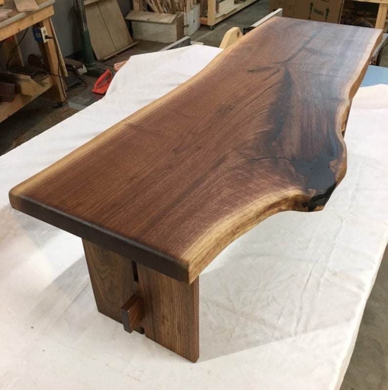 May include: A dark brown live edge walnut wood coffee table with a natural edge and a clear epoxy fill. The table has a simple, modern design with two legs made of the same wood.