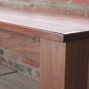 May include: A close-up of a wooden bench with a smooth, dark brown finish. The bench has a simple, rectangular design with straight legs and a wide, flat top.
