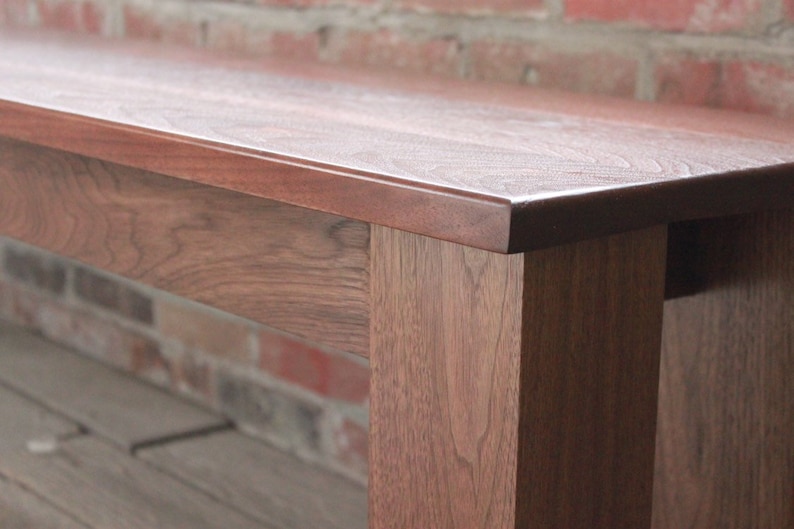 May include: A close-up of a wooden bench with a smooth, dark brown finish. The bench has a simple, rectangular design with straight legs and a wide, flat top.