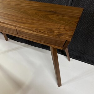 May include: A rectangular wooden console table featuring a rich brown grain pattern, a smooth flat top surface, and two integrated drawers positioned horizontally. The furniture piece is supported by four tapered legs angled outward from the base.