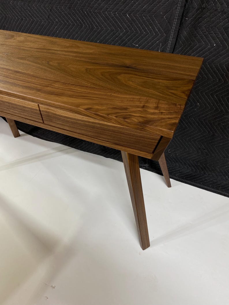May include: A rectangular wooden console table featuring a rich brown grain pattern, a smooth flat top surface, and two integrated drawers positioned horizontally. The furniture piece is supported by four tapered legs angled outward from the base.