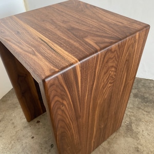 May include: A solid wood side table with a square top and angled legs. The table is made of dark brown wood with a visible grain pattern. The design is modern and minimalist, suitable for various interior styles.