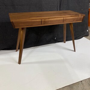 May include: A rectangular wooden desk crafted from dark brown walnut wood, featuring a smooth flat top surface and three horizontal drawers aligned across the front. The desk is supported by four tapered, angled legs extending outward.
