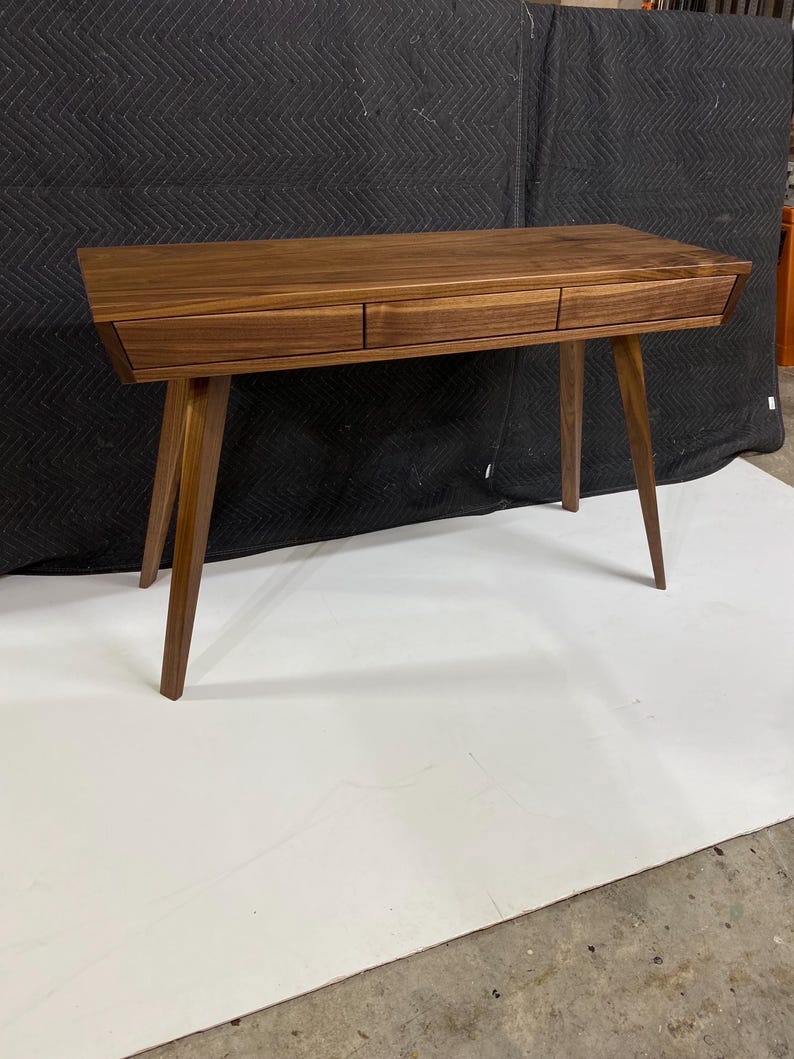 May include: A rectangular wooden desk crafted from dark brown walnut wood, featuring a smooth flat top surface and three horizontal drawers aligned across the front. The desk is supported by four tapered, angled legs extending outward.