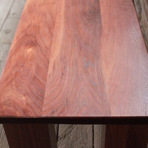 May include: A close-up of a wooden bench with a smooth, reddish-brown finish. The grain of the wood is visible and the bench has a simple, rectangular design.