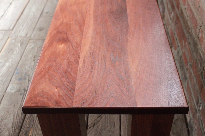 May include: A close-up of a wooden bench with a smooth, reddish-brown finish. The grain of the wood is visible and the bench has a simple, rectangular design.