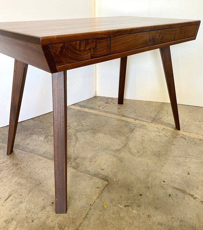 May include: A dark brown wooden desk with a single drawer and four legs. The desk is angled to the left and the legs are slightly flared at the bottom.
