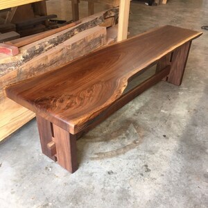 May include: A long, wooden bench made from a single piece of walnut with a live edge. The bench has a simple, modern design with two legs on each end. The bench is made of dark brown wood with a natural finish.