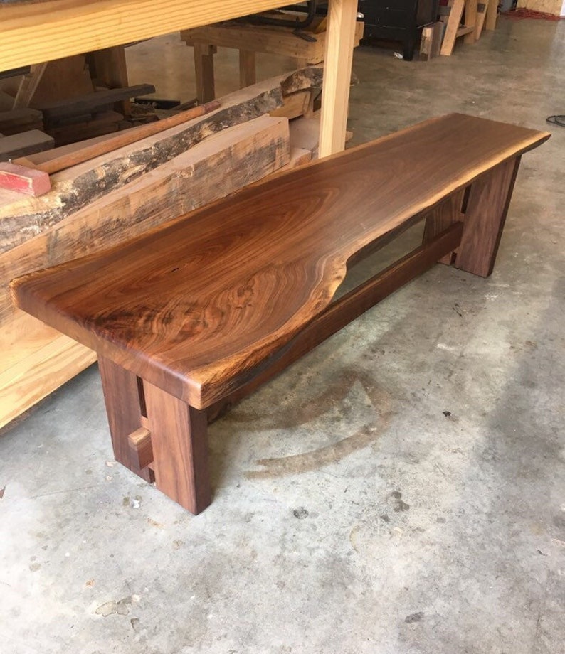 May include: A long, wooden bench made from a single piece of walnut with a live edge. The bench has a simple, modern design with two legs on each end. The bench is made of dark brown wood with a natural finish.