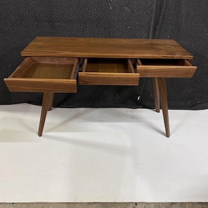 May include: A rectangular wooden desk crafted from dark walnut wood with a smooth grain finish. The desk features three open drawers positioned horizontally across the front and is supported by four angled, tapered wooden legs.