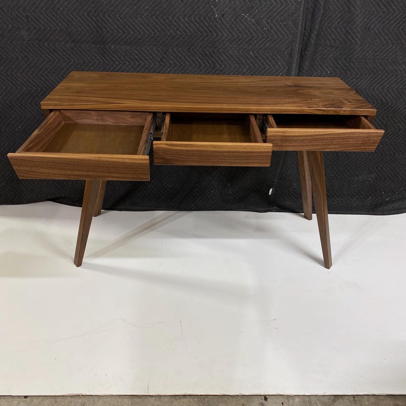 May include: A rectangular wooden desk crafted from dark walnut wood with a smooth grain finish. The desk features three open drawers positioned horizontally across the front and is supported by four angled, tapered wooden legs.