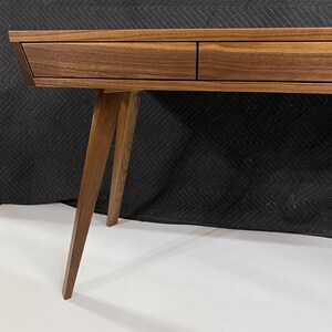 May include: A brown wooden desk featuring a smooth, flat top surface and two integrated drawers positioned side by side. The desk is supported by angled, tapered wooden legs that extend outward from the main frame, showcasing a natural wood grain.
