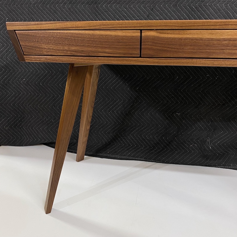 May include: A brown wooden desk featuring a smooth, flat top surface and two integrated drawers positioned side by side. The desk is supported by angled, tapered wooden legs that extend outward from the main frame, showcasing a natural wood grain.