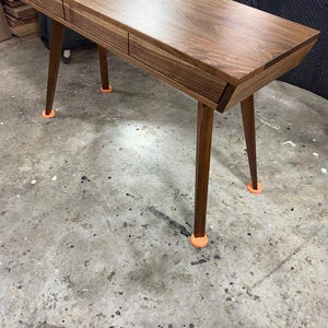 May include: A rectangular wooden desk crafted from dark walnut wood featuring a smooth, polished surface and two integrated drawers. The desk is supported by four tapered, angled legs, each resting on a small, bright orange circular floor protector.