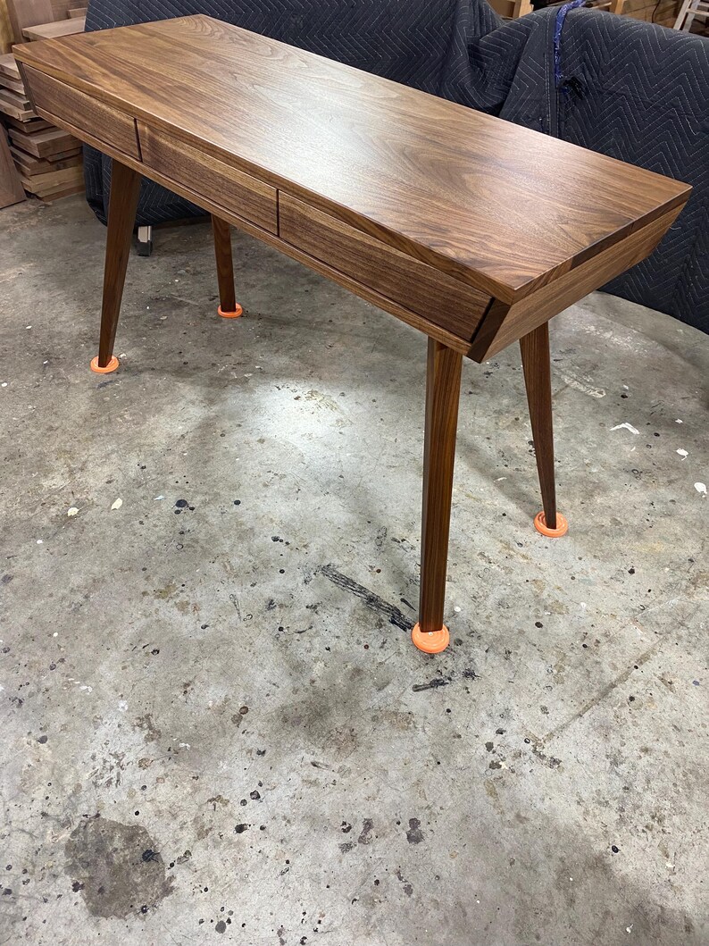 May include: A rectangular wooden desk crafted from dark walnut wood featuring a smooth, polished surface and two integrated drawers. The desk is supported by four tapered, angled legs, each resting on a small, bright orange circular floor protector.