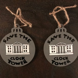 May include: Two gray Christmas ornaments with black and white lettering. The ornaments are round and have a string attached to the top. The text on the ornaments reads "Save the Clock Tower".