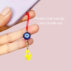 May include: A red string with a blue evil eye charm and a gold hamsa hand charm. The text "Protect your energy. Carry it with you." is written in white on a pink background. The charms are attached to a phone case.