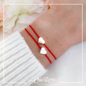 May include: Two red string bracelets, each featuring a small, polished silver-tone heart charm. The bracelets are worn on a wrist, with the text ChicLove ACCESSORIES visible at the bottom of the frame in a white script font.