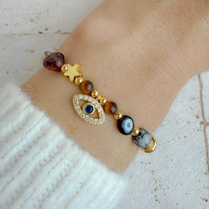 May include: A gold-toned bracelet featuring an evil eye charm with a blue center stone, a gold star, and various beads in brown, gold, and purple hues. The bracelet is worn on a wrist, with a white sweater sleeve in the background.